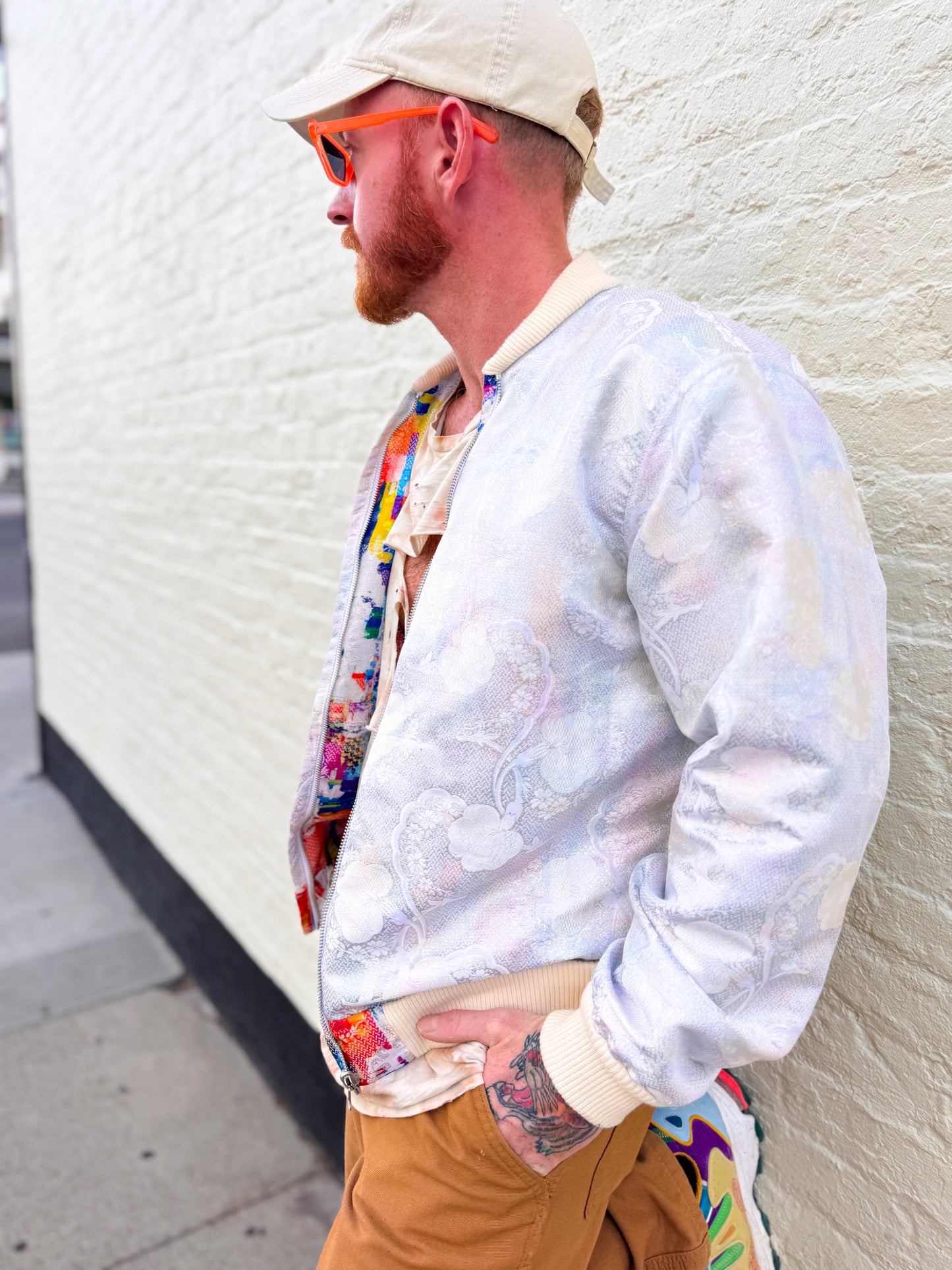 Man wearing a light-colored jacket, colorful shirt, and beige cap against a white wall.