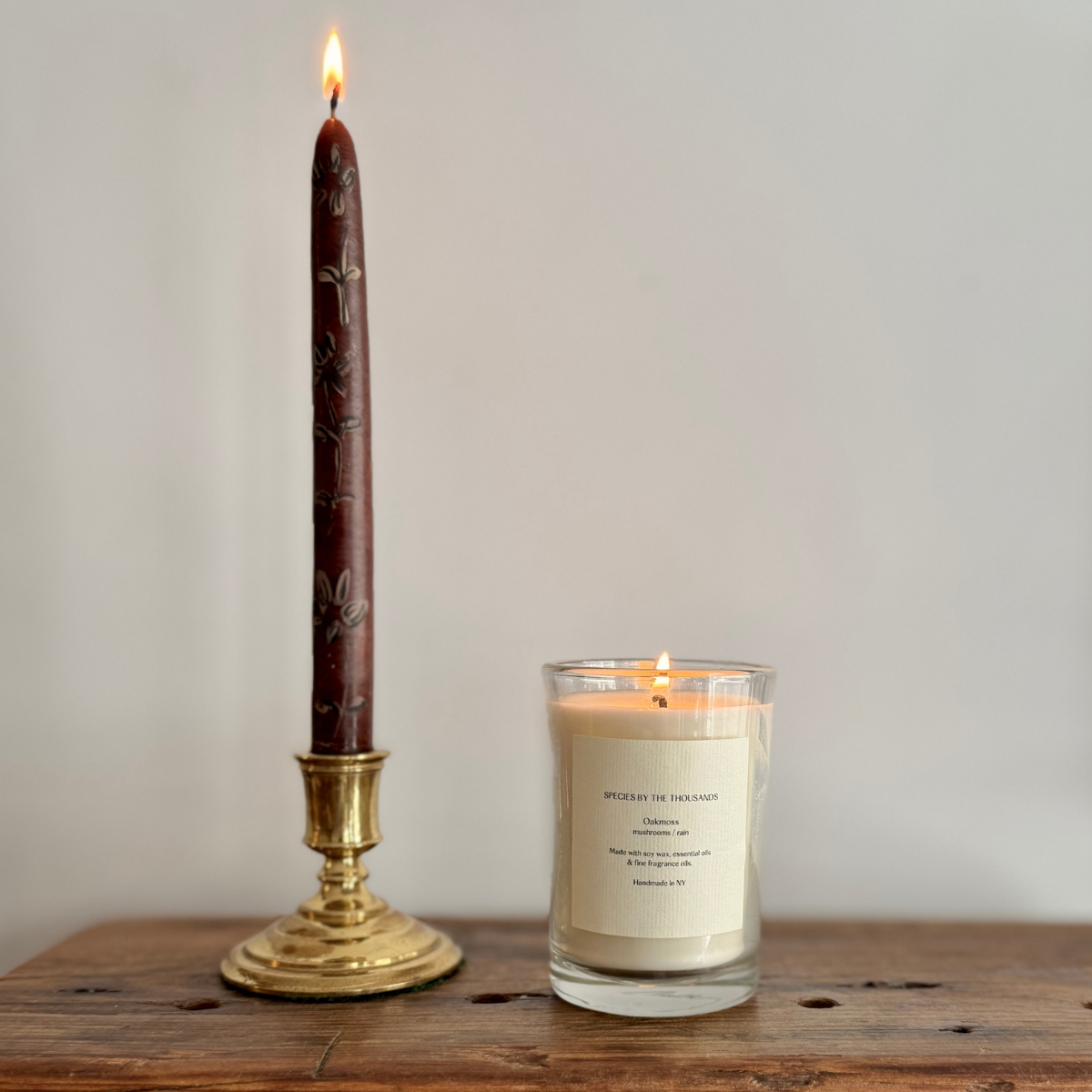 Lit candle in glass next to lit taper candle, sitting atop a wood surface.