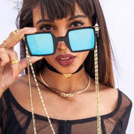 Woman holding sunglasses with a gold chain, wearing a black sheer top and gold jewelry.