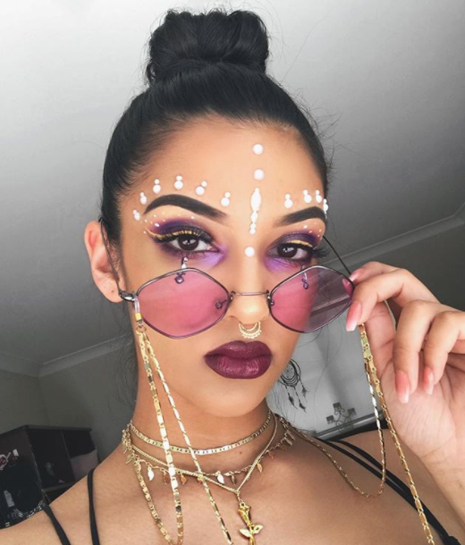 Person wearing pink sunglasses with chain details, dark makeup, and jewelry indoors.