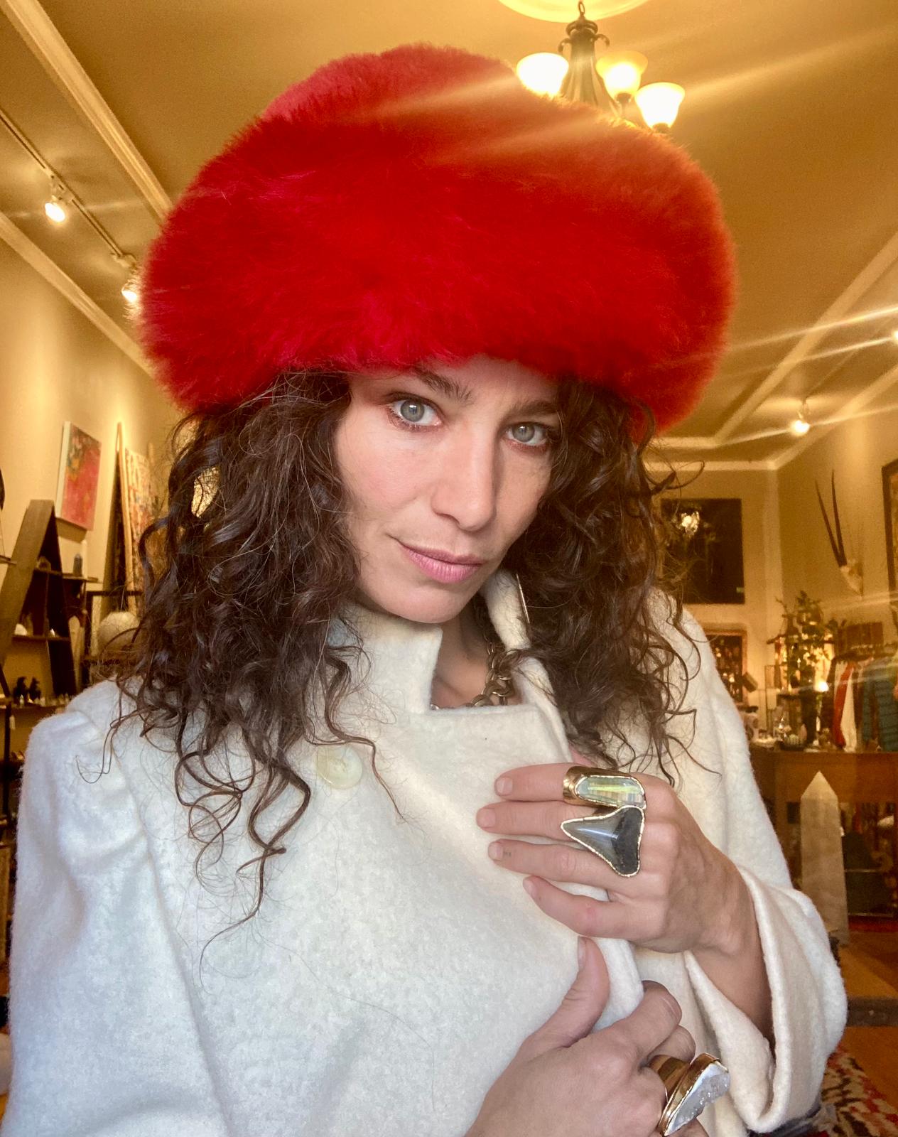 A person wearing a red Alpaca fur hat and a white coat, holding a ring, with various items displayed in the background.