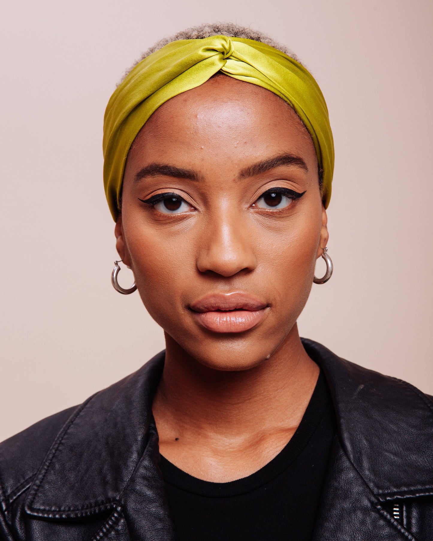 Woman wearing a yellow headscarf and black leather jacket against a beige background