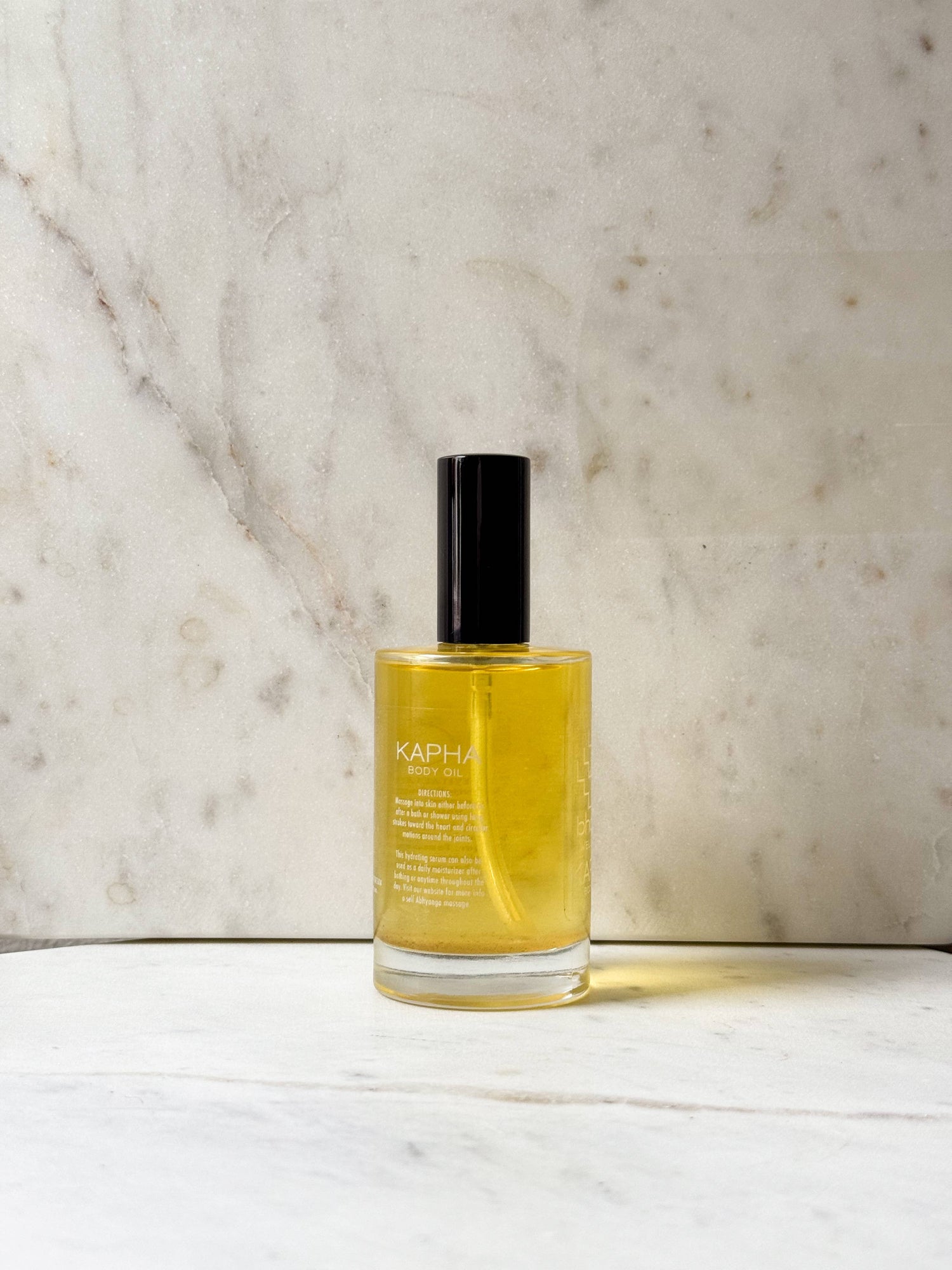 A clear glass bottle of KAPHA body oil filled with golden yellow liquid, featuring a matte black cap, set against a light beige marble background.