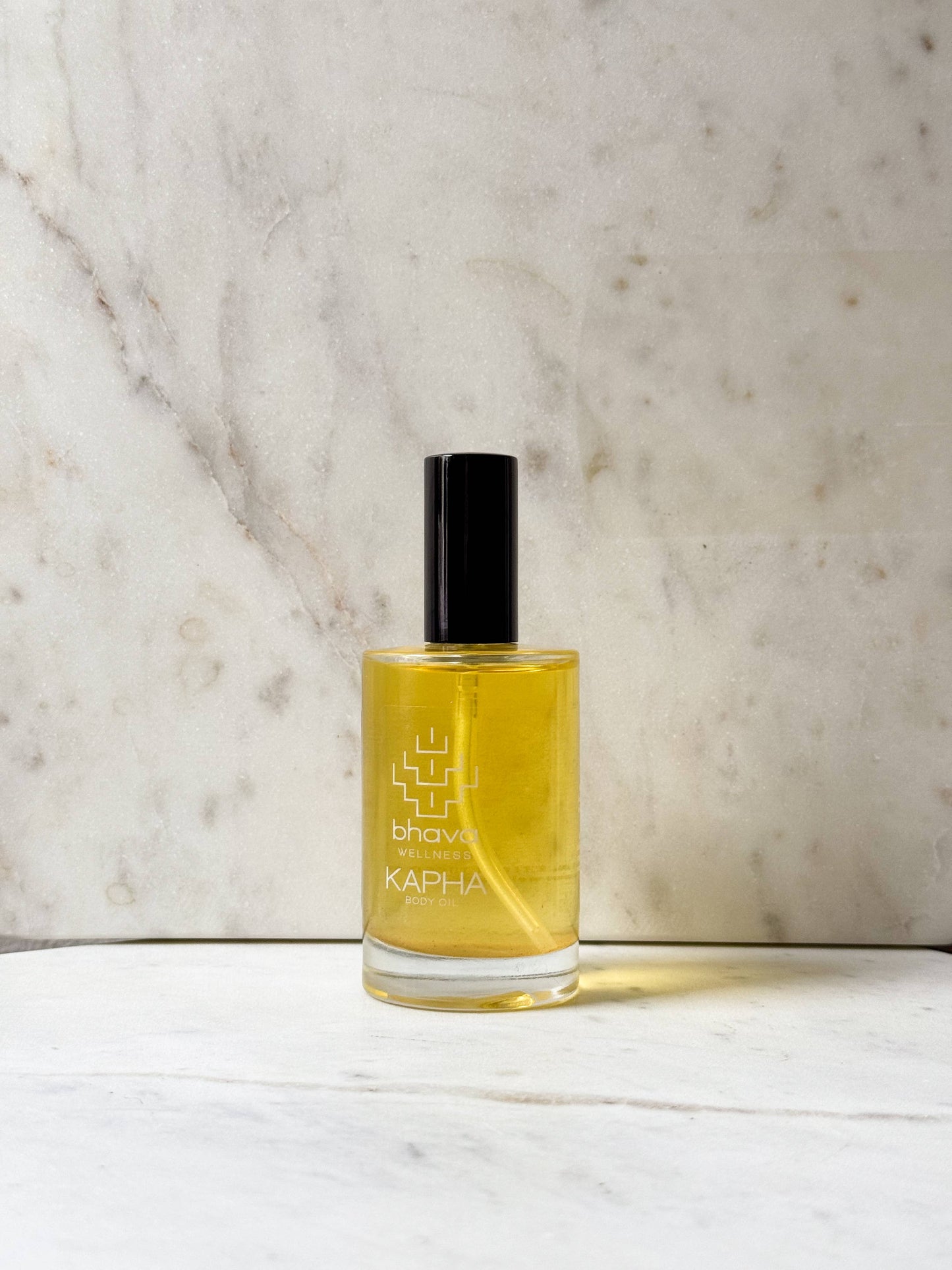 A bottle of Kapha body oil by Bhavi Smiles, filled with golden yellow oil and featuring a black cap, set against a light marble background.