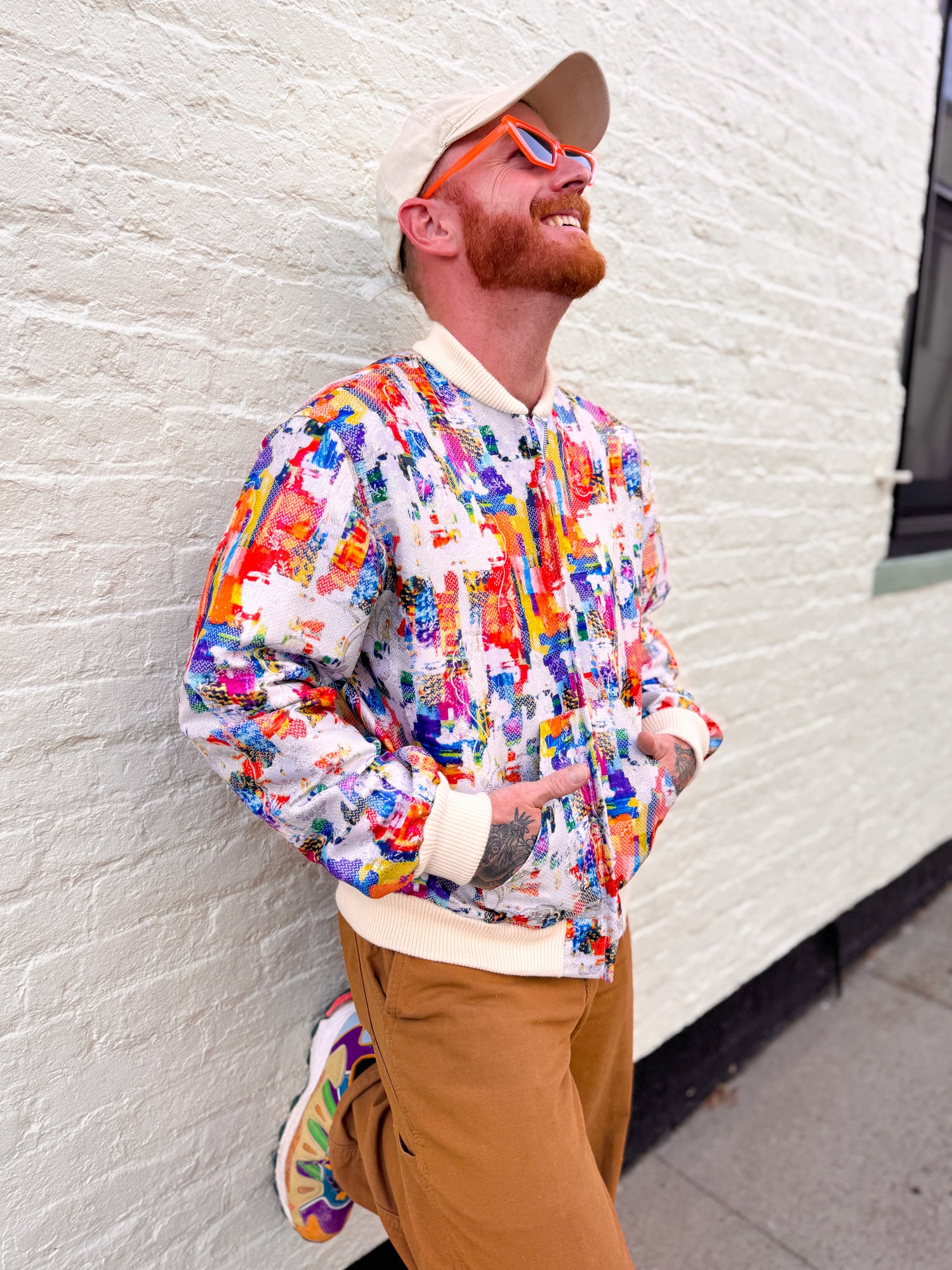 Man wearing a colorful jacket and brown pants leaning against a white wall.