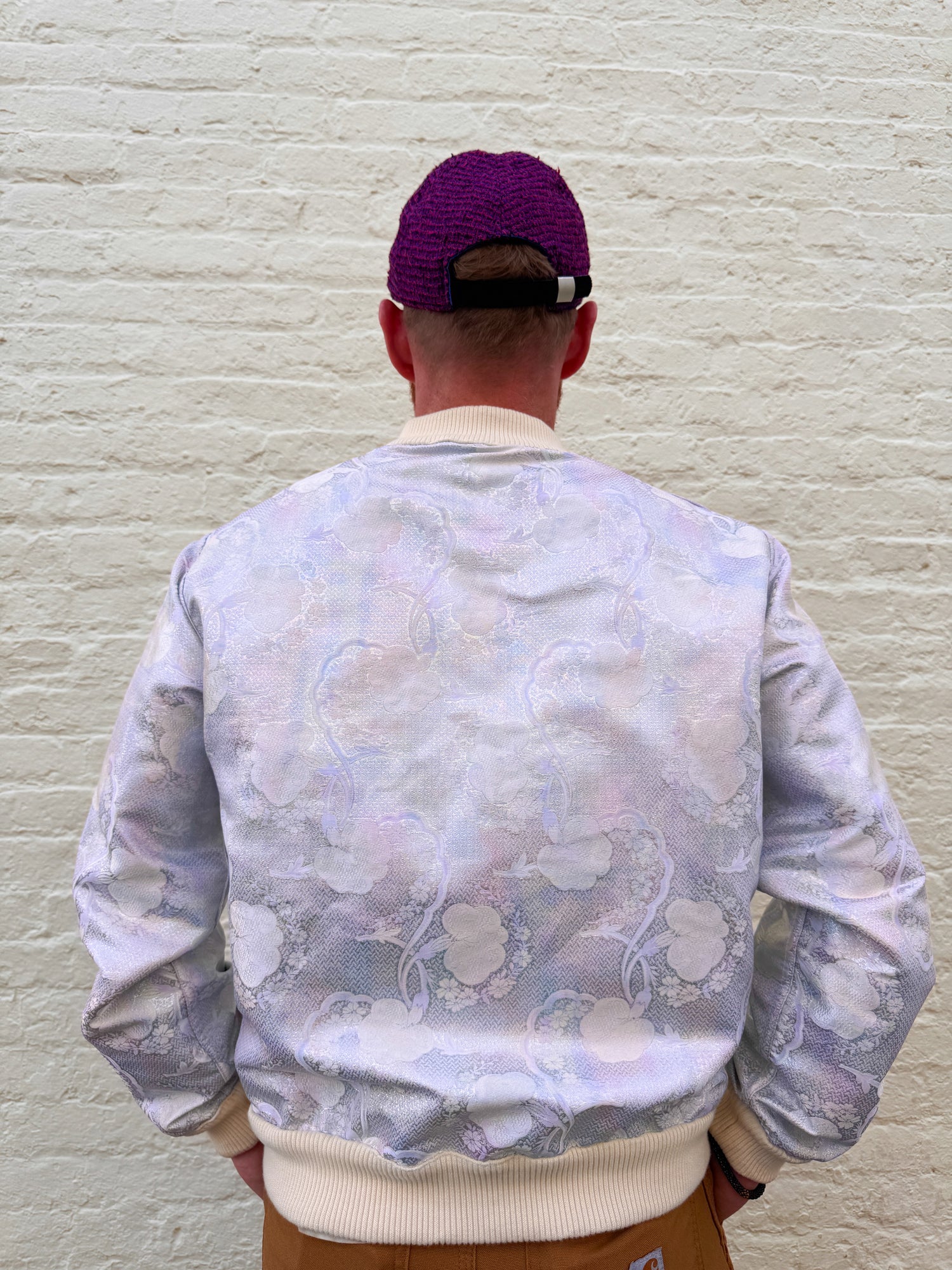 Person wearing a floral-patterned bomber jacket and purple cap against a textured white wall.