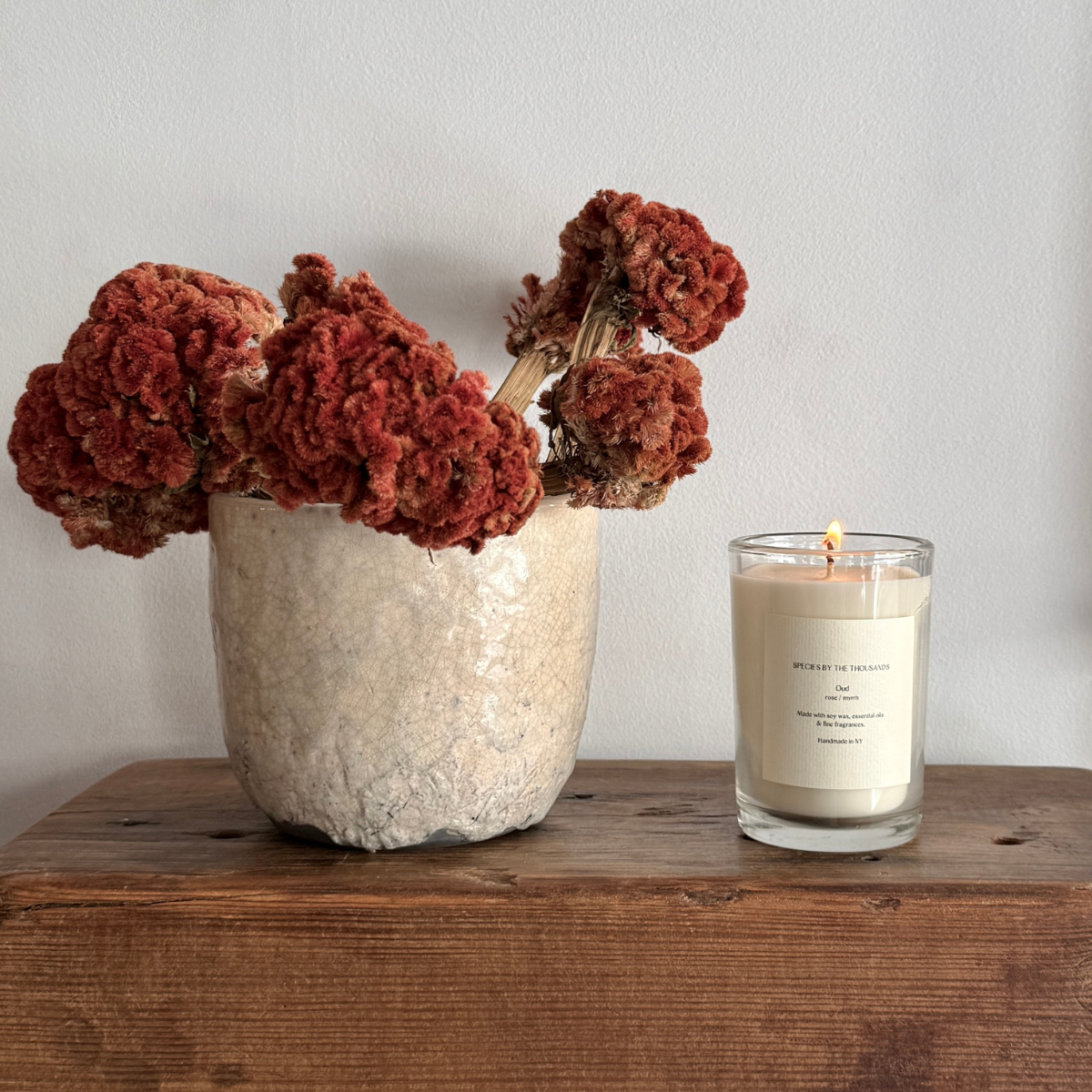 Lit candle in glass jar sat atop a wood table next to red-orange flowers in a cream-colored earthen pot.