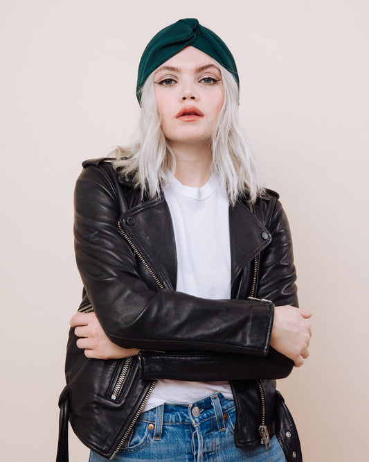 Person wearing a black leather jacket, white shirt, blue jeans, and green headscarf against a beige background