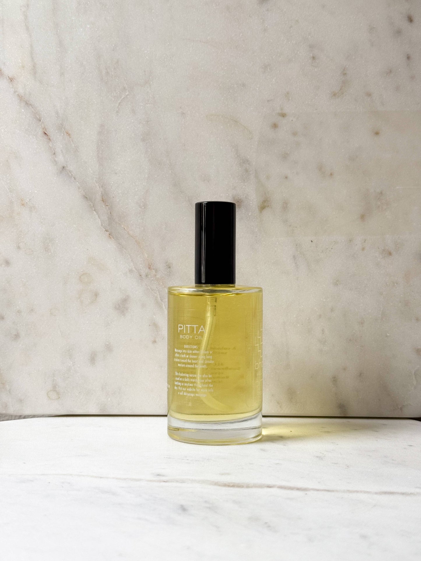 A clear glass bottle of Pitta London body mist with pale yellow liquid and a black cap, standing on a white marble surface against a beige marble background.
