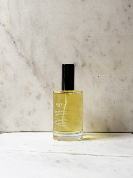 A clear glass bottle of Pitta London body mist with pale yellow liquid and a black cap, standing on a white marble surface against a beige marble background.