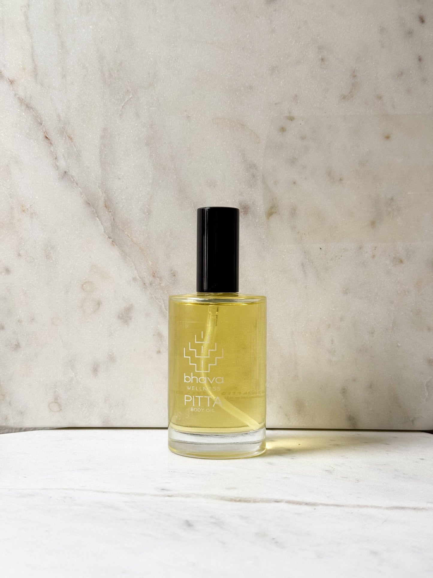 A bottle of bhava aromas PITHA essential oil or perfume with pale yellow liquid, a black cap, and a clear glass base, set on a white marble surface against a beige marble background.