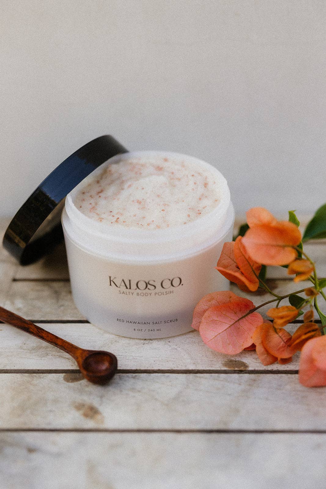 Jar of Kalos Co. body scrub on a wooden surface with flowers and a spoon.