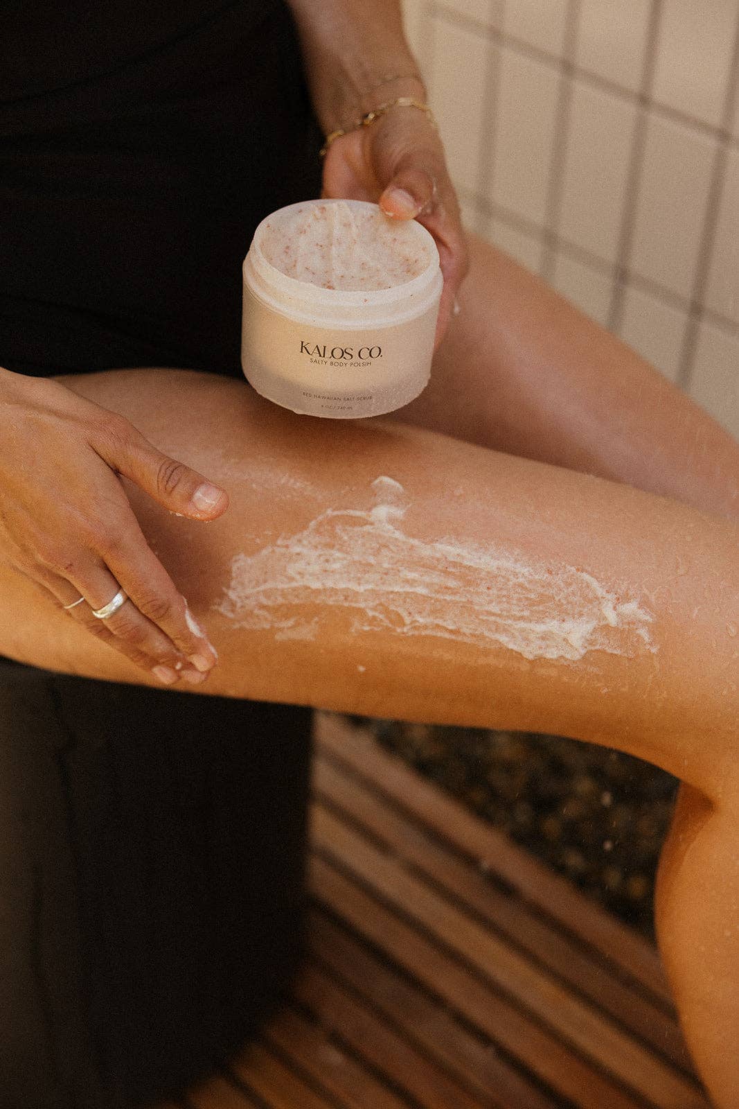 Person applying salty body scrub to their leg with a jar of the product in hand.