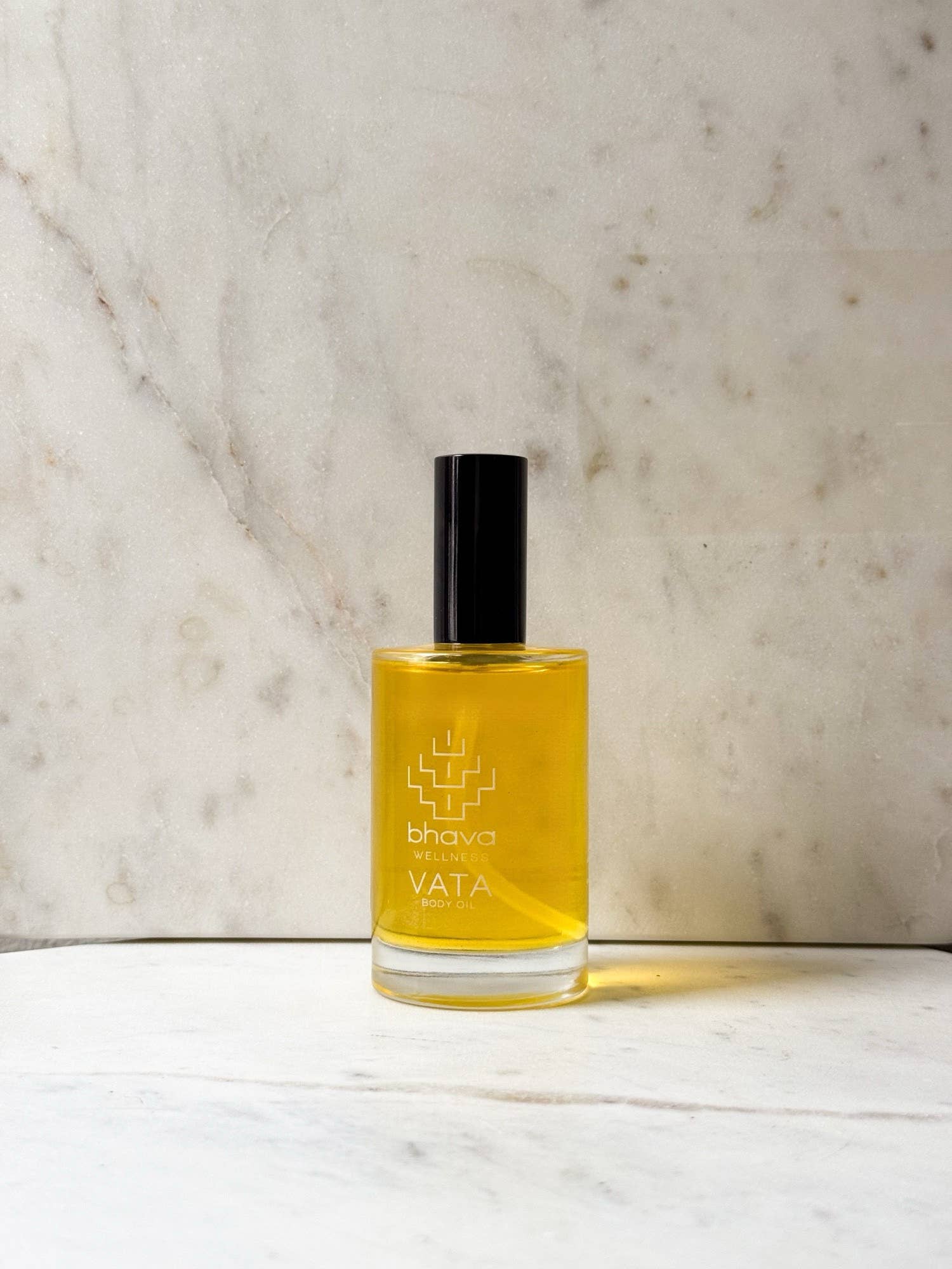 A bottle of Bhava Vata Ayurvedic oil with golden yellow liquid, a black cap, and white text, set on a white marble surface against a beige marble background.