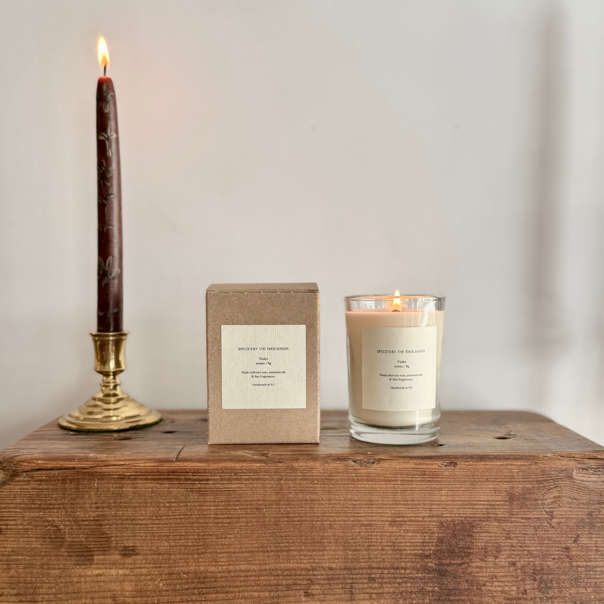 Lit candle in glass jar sat atop a wood table next to a sand-colored box and a tall, lit taper candle in a gold candlestick holder.